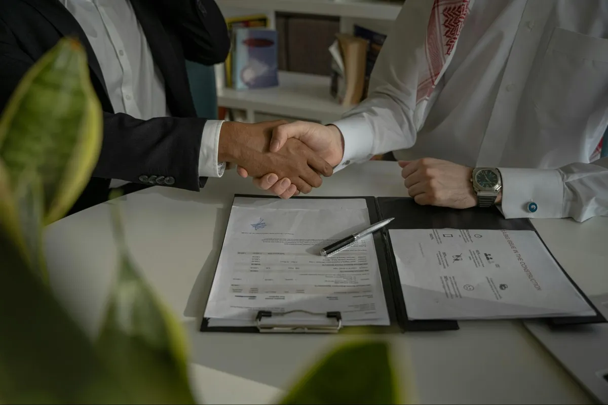 Zwei Personen geben sich über einem Vertrag und einem Stift auf einem Bürotisch die Hand